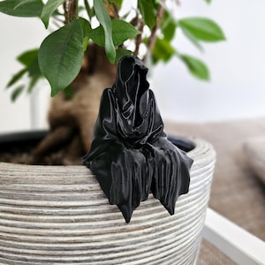 May include: A black figurine of a hooded figure sitting on the edge of a white planter. The figurine is made of a smooth, matte material and has a detailed design.
