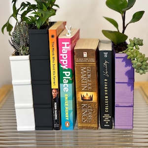 Może przedstawiać: Dekoracyjna aranżacja z książkami i roślinami. Książki, z tytułami takimi jak "Happy Place" i "Verity", są przeplatane sukulentami i inną zielenią. Donice są białe, czarne i fioletowe, tworząc kolorową ekspozycję.