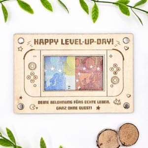 May include: A wooden gift card holder shaped like a video game console, with the text "HAPPY LEVEL-UP-DAY!". The center displays Euro banknotes. The bottom text reads "DEINE BELOHNUNG FÜRS ECHTE LEBEN. GANZ OHNE QUEST!".