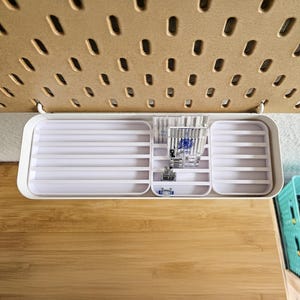 May include: A white plastic organizer with multiple compartments, hanging from a pegboard. The organizer contains sewing machine needles, presser feet, and other sewing notions. The pegboard is a light brown color.