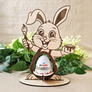 May include: Wooden Easter bunny egg holder with a paintbrush and a cup. The bunny is holding a Kinder Surprise egg.