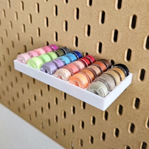 Può includere: Una mensola bianca contiene una collezione di rocchetti di filo colorati. Le rocchetti sono disposti in fila, mostrando una varietà di colori tra cui rosa, verde, blu, rosso, arancione e marrone. La mensola è montata su una pannello forato.