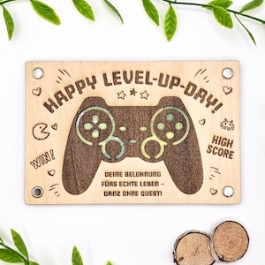 May include: A wooden plaque with the words "HAPPY LEVEL-UP-DAY!" and a controller design. The plaque includes the words "WIN!" and "HIGH SCORE". The text "DEINE BELOHNUNG FÜRS ECHTE LEBEN - GANZ OHNE QUEST!" is also present.