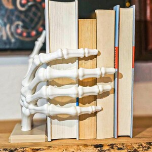 May include: A white, skeletal hand bookend holds several books on a wooden shelf. The bookend is detailed with bone-like textures, and the books have varying colors and sizes, creating a unique decorative piece for a bookshelf.