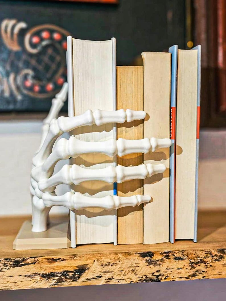 May include: A white, skeletal hand bookend holds several books on a wooden shelf. The bookend is detailed with bone-like textures, and the books have varying colors and sizes, creating a unique decorative piece for a bookshelf.