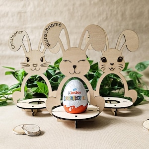 May include: Three wooden bunny-shaped Easter egg holders. The center bunny has a chocolate egg inside. The bunnies are personalized with the names "Christopher", "Wunschname", and a blank space for a third name.