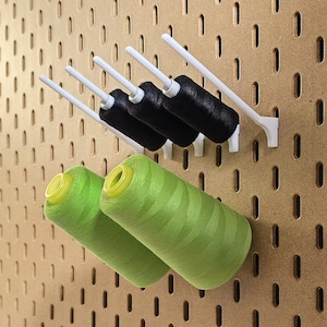 May include: A pegboard with white thread spool holders and spools of black and green thread. The green thread spools have yellow centers. The pegboard is made of a light brown material.
