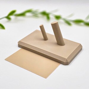 May include: A beige-colored, rectangular display stand with two cylindrical pegs and a matching rectangular card. The stand is made of a solid material and is designed to hold or display small items. The background is white with a blurred green plant.