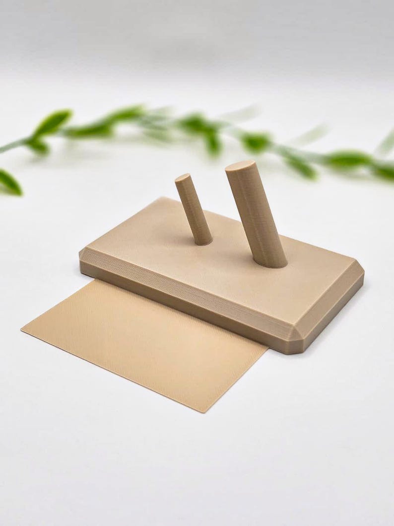May include: A beige-colored, rectangular display stand with two cylindrical pegs and a matching rectangular card. The stand is made of a solid material and is designed to hold or display small items. The background is white with a blurred green plant.