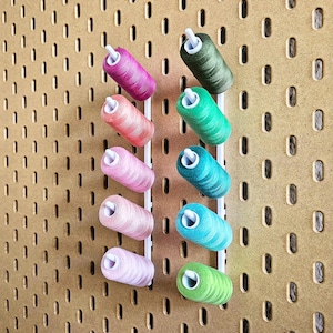 May include: A selection of colourful thread spools in shades of pink, green, and blue, arranged on a pegboard. The spools are held in place by white plastic holders, creating a visually appealing display for sewing or crafting.