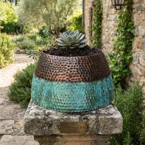 May include: A hammered metal planter with a two-tone finish, featuring a dark copper upper section and a vibrant turquoise patina lower section. The bowl-shaped vessel contains a green succulent plant and sits on a stone pillar with a small plaque.