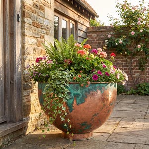 May include: A large, round, bowl-shaped planter made of copper with a distinct verdigris patina finish. The planter is filled with a lush arrangement of green ferns, trailing ivy, and various colourful flowers in shades of pink, red, and orange.