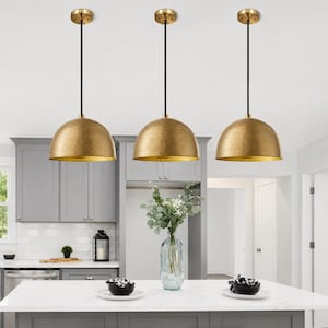 May include: Three dome-shaped pendant lights with a textured, hammered gold finish. Each light features a thin black cord and a circular gold ceiling canopy. The fixtures are suspended in a row, showcasing a metallic surface and a rounded shape.