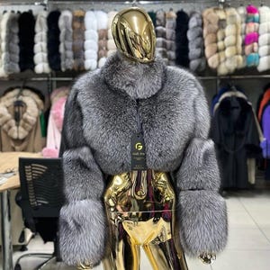 Puede incluir: Un bolero de piel de color gris plateado con cuello alto y mangas largas, exhibido en un maniquí dorado. La chaqueta tiene un aspecto lujoso y texturizado. El fondo muestra una variedad de abrigos de piel.