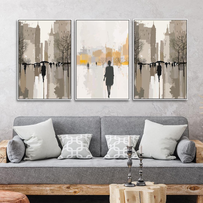 May include: Set of three framed abstract art prints. Each print features a cityscape with silhouetted figures walking in the rain. The color palette includes shades of gray, brown, and yellow, creating a moody, urban scene.