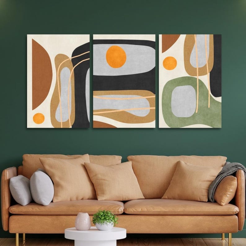 May include: Triptych wall art featuring abstract shapes in brown, black, gray, and green with orange circles. The artwork is displayed above a brown leather sofa with tan pillows and a white side table. The wall behind the sofa is painted a dark green.