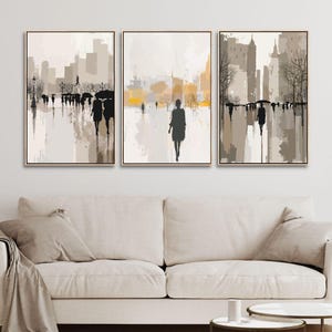 May include: Triptych of abstract cityscape art prints. Each framed canvas features a muted color palette of beige, gray, and yellow, depicting silhouetted figures walking on a wet street with buildings in the background. The artwork is displayed above a beige sofa.