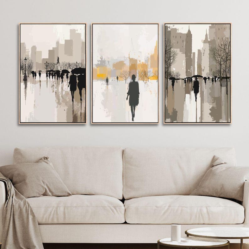 May include: Triptych of abstract cityscape art prints. Each framed canvas features a muted color palette of beige, gray, and yellow, depicting silhouetted figures walking on a wet street with buildings in the background. The artwork is displayed above a beige sofa.