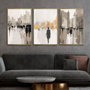 May include: Set of three framed abstract art prints. Each print features a muted color palette of beige, gray, and gold, depicting figures walking in a city setting. The artwork is displayed above a dark gray sofa.