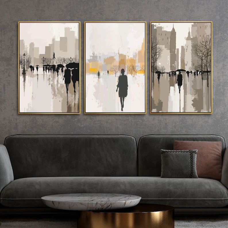 May include: Set of three framed abstract art prints. Each print features a muted color palette of beige, gray, and gold, depicting figures walking in a city setting. The artwork is displayed above a dark gray sofa.