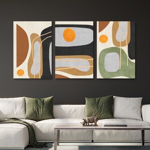 May include: Triptych wall art featuring abstract shapes in earth tones and black against a cream background. Each panel has a unique composition with orange circles and linear accents. The artwork is displayed above a white sofa with green pillows.