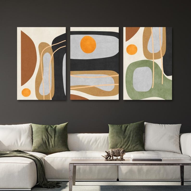 May include: Triptych wall art featuring abstract shapes in earth tones and black against a cream background. Each panel has a unique composition with orange circles and linear accents. The artwork is displayed above a white sofa with green pillows.