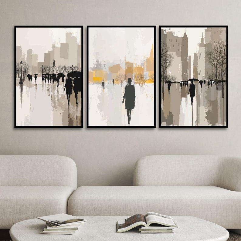 May include: Set of three framed art prints depicting an urban scene with silhouetted figures walking in the rain. The artwork features a muted color palette of beige, gray, and yellow, with a modern, abstract style. The prints are displayed above a white sofa.