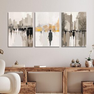 May include: A triptych of abstract cityscape paintings. Each canvas features a muted color palette of beige, gray, and black, depicting silhouetted figures walking in a city setting. The artwork evokes a rainy day atmosphere with reflections.