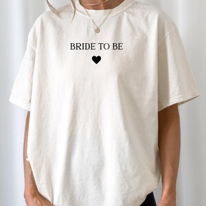Puede incluir: Camiseta color crema con el texto negro "BRIDE TO BE" y un símbolo de corazón negro. La camiseta es de corte holgado, con mangas cortas. La modelo lleva un pantalón corto negro.