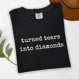 May include: A black short-sleeved t-shirt featuring the white text "turned tears into diamonds" printed in a typewriter-style font across the chest. The garment has a crew neckline and a Comfort Colors brand label visible at the inner collar.
