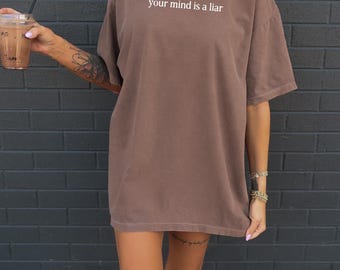 May include: A brown t-shirt with the text "your mind is a liar" in white. The shirt is oversized and appears to be made of a soft material. The person is holding a drink with a straw.
