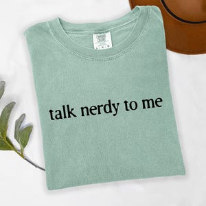 Peut inclure: Un t-shirt vert clair avec l'inscription "talk nerdy to me" en noir. Le t-shirt a un col rond et est fait d'une matière douce. Un chapeau marron est visible en arrière-plan.