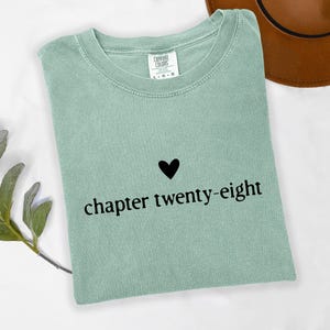 May include: A sage green t-shirt with the words "chapter twenty-eight" in black font, with a black heart above the text. The shirt is folded and has a tag at the neckline. A brown hat is visible in the background.