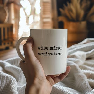 Könnte beinhalten: Eine weiße Keramiktasse mit den schwarzen Aufdruck "wise mind activated". Die Tasse wird von einer Hand gehalten, mit einem verschwommenen Hintergrund eines gemütlichen Interieurs. Die Tasse hat eine Standardgröße, ideal für Kaffee oder Tee.
