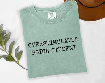 May include: A pale green t-shirt with the text "OVERSTIMULATED PSYCH STUDENT" in black. The shirt is folded and has a crew neck. A brown hat is visible in the background.