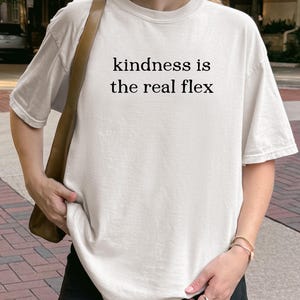 May include: Off-white t-shirt with the text "kindness is the real flex" in black. The shirt is a casual style, with short sleeves and a relaxed fit. The person is wearing a black skirt and carrying a brown shoulder bag.