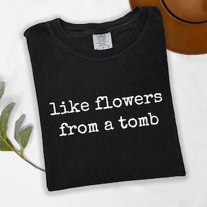 May include: A black short-sleeved t-shirt featuring the white text "like flowers from a tomb" printed in a typewriter-style font across the chest. The garment has a crew neckline and a visible brand label on the inside of the collar.