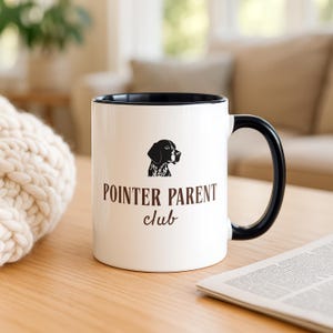 May include: White ceramic mug with a black interior and handle. The mug features a black silhouette of a dog's head above the words "POINTER PARENT club" in brown lettering. The mug is on a wooden table.