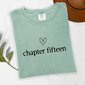 May include: A light green Comfort Colors t-shirt with the words "chapter fifteen" in black script below a black heart. The shirt is folded and lies on a white surface.