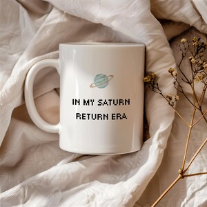 May include: White ceramic mug with a handle, featuring a light blue and gold planet graphic and the text "IN MY SATURN RETURN ERA" in black. The mug is set against a neutral, textured background.