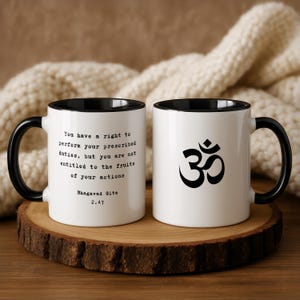 May include: Two white ceramic mugs with black rims and handles. One mug has the text "You have a right to perform your prescribed duties, but you are not entitled to the fruits of your actions" and "Bhagavad Gita 2.47". The other mug has an Om symbol.