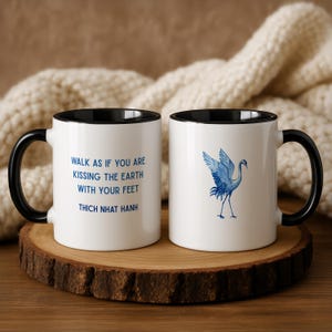 Peut inclure: Deux mugs en céramique blancs avec des anses et des bords noirs. L'un des mugs présente le texte "WALK AS IF YOU ARE KISSING THE EARTH WITH YOUR FEET THICH NHAT HANH" en bleu. L'autre mug a une illustration de grue bleue.