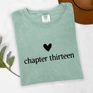 May include: A sage green t-shirt with a black heart graphic above the words "chapter thirteen". The shirt is folded and lies on a white surface, with a brown hat visible in the background.