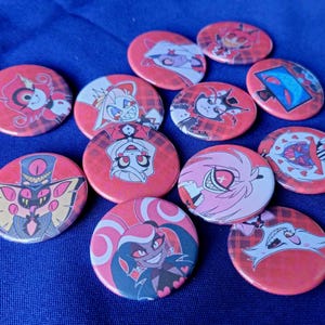 May include: A set of 12 round red and white buttons featuring cartoon characters from the animated series "Hazbin Hotel".