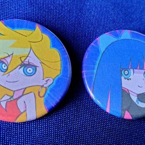 May include: Two round anime character buttons. The first button features a blonde character with blue eyes wearing a red dress. The second button features a blue-haired character with blue eyes wearing a black dress.