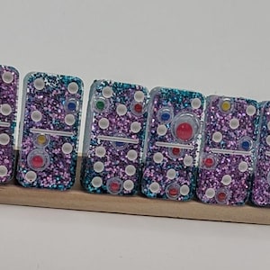 Op de afbeelding: Zes met glitter bedekte dominostenen in een houten houder. De dominostenen hebben een paarse en blauwgroene glitterbasis met kleurrijke stippen. De stippen zijn rood, geel, groen en blauw. De dominostenen zijn in een rij gerangschikt.