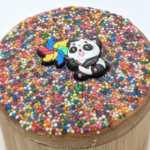 Könnte beinhalten: Ein kleiner, runder Holzbehälter mit einer bunten, mit Streuseln bedeckten Oberseite. Auf der Oberseite befindet sich eine Panda-Bären-Figur mit einem Regenbogen-Windrad. Der Behälter besteht aus hellbraunem Holz.