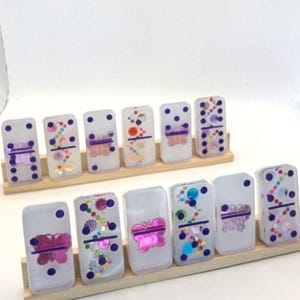 Puede incluir: Un juego de dominó blanco con diseños de mariposas moradas y rosadas y detalles de purpurina de colores. Los dominós están dispuestos en dos filas sobre un soporte de madera.