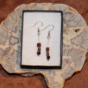 May include: A pair of silver earrings with brown gemstone beads. The earrings are displayed in a white box with a black border.