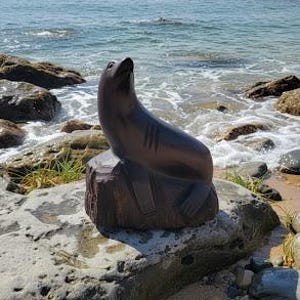 Op de afbeelding: Een bruine houten sculptuur van een zeehond, geplaatst op een rots bij de oceaan. De zeehond is afgebeeld in een ontspannen pose, met zijn hoofd omhoog gekanteld. De sculptuur staat tegen een achtergrond van zee en lucht.